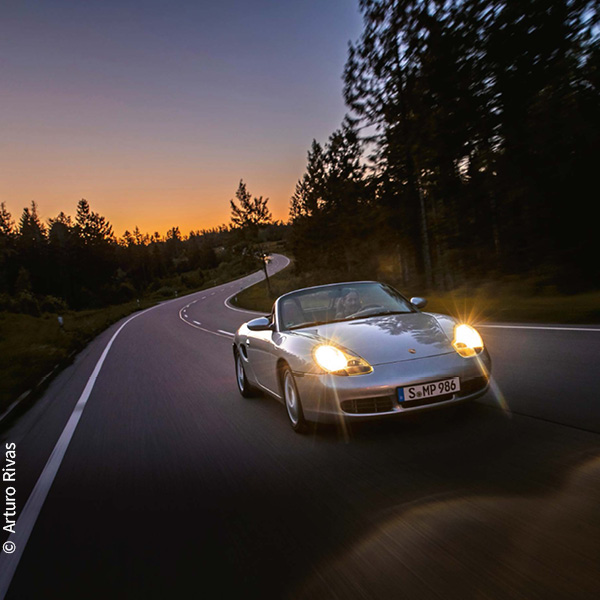ein silberfarbener Porsche Boxster S fährt mit eingeschalteten Scheinwerfern in der Dämmerung eine Straße entlang