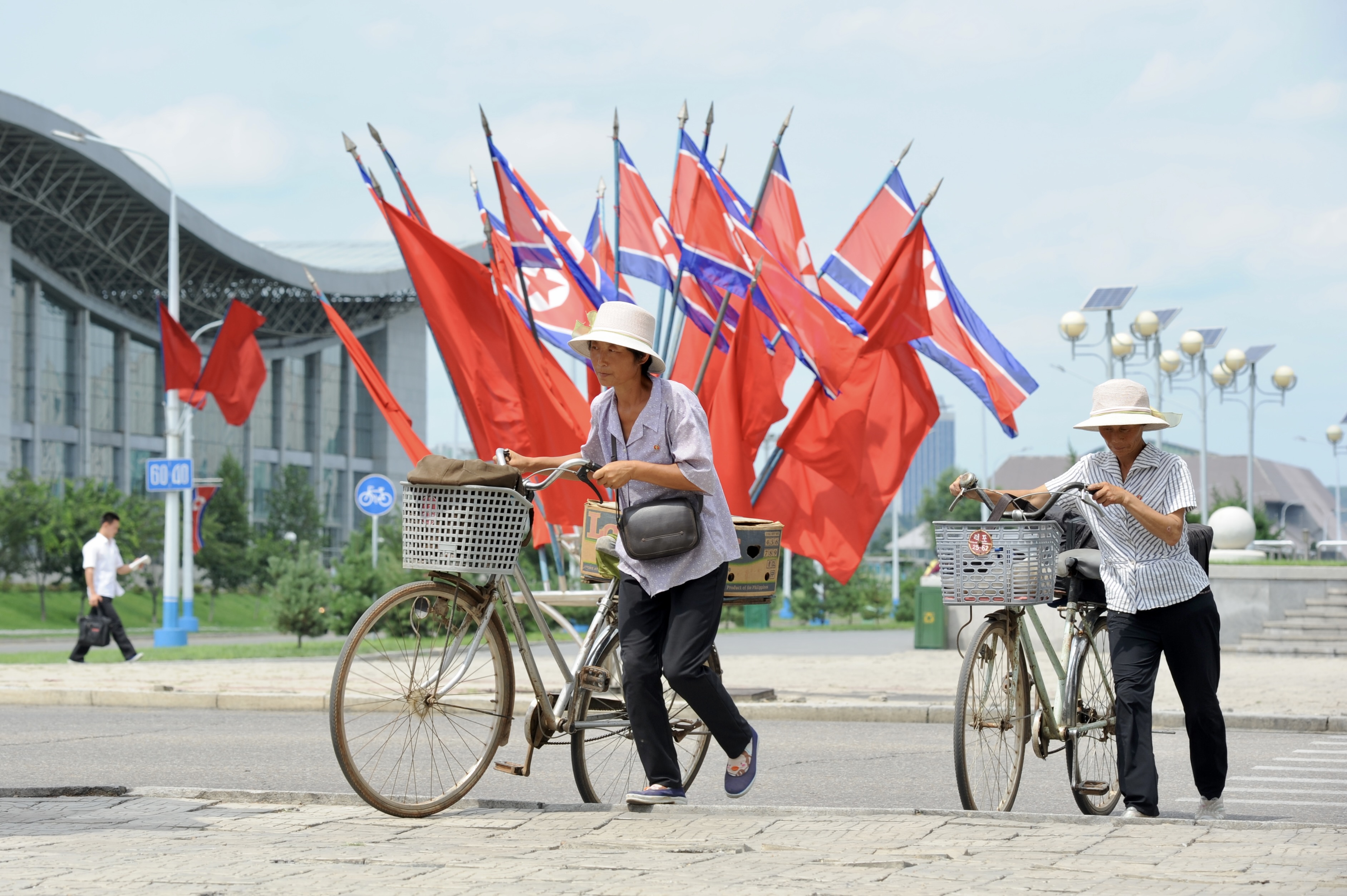 Sommerliche Szene aus der reichlich mit Flaggen geschm&uuml;ckten Hauptstadt: Zwei Frauen schieben ihre Fahrr&auml;der ueber einen Zebrastreifen. Im Hintergrund monumentale Architektur und Fahnenschmuck, sichtbare Zeichen der politischen Dimension des Landes.